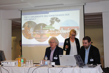 2 Personen und eine Rednerin auf Podium vor Präsentation