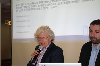 ein Redner mit weiterer Person auf Podium vor Präsentation