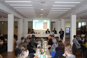 Teilnehmer Arbeitskreis an langer Tafel mit Blick zum Podium vor Präsentation